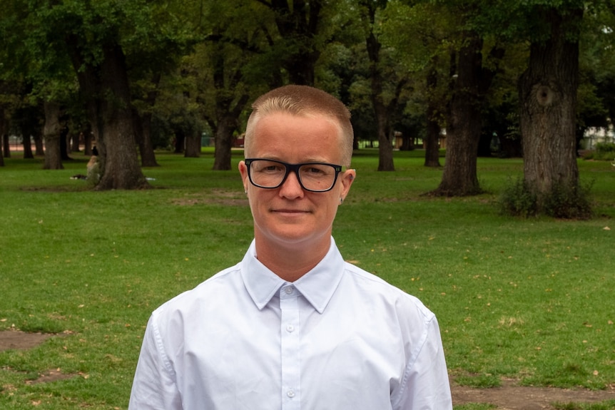 A headshot of a non-binary person in a white shirt wearing black-framed glasses taken in a park