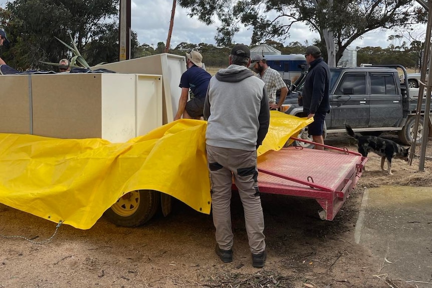 Four men load a white wardrobe onto the back of a trailer that has a yellow tarp on it
