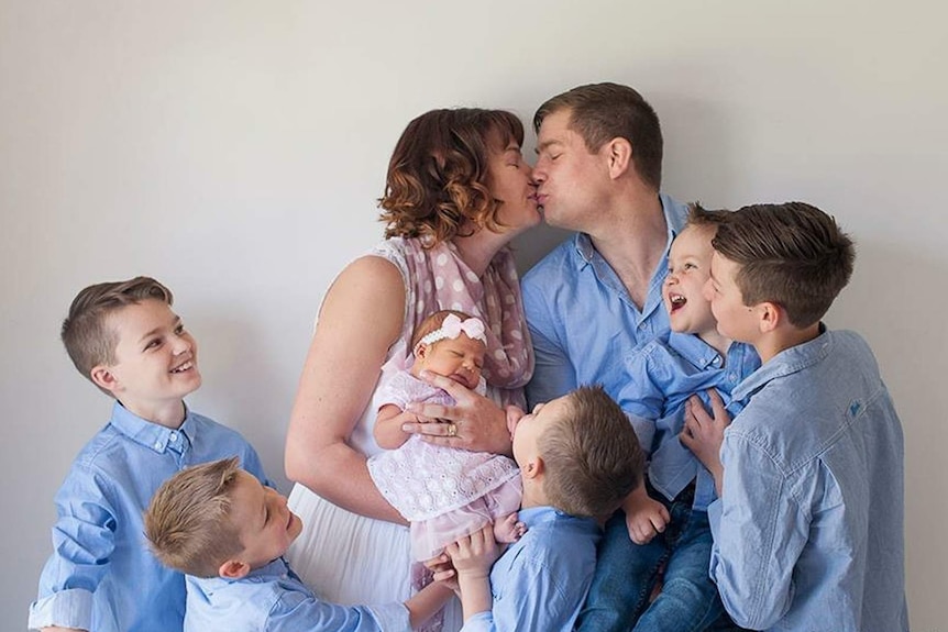A man kissing his wife who is holding a baby, surrounded by six boys