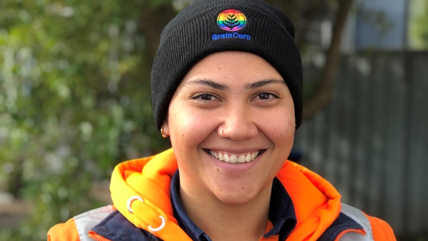 A smiling maori woman with short hair wears a black beanie with a rainbow grain corp logo on it and orange hi vis jacket