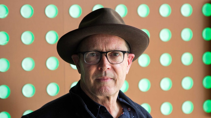 A man in a hat and glasses looks at the camera, green circular lights in background.