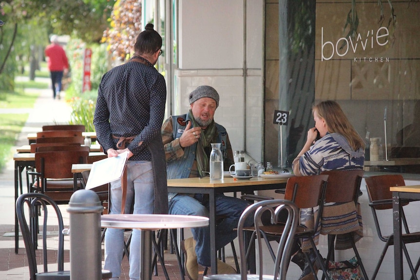 A man and woman sit at a cafe speaking to a man standing up