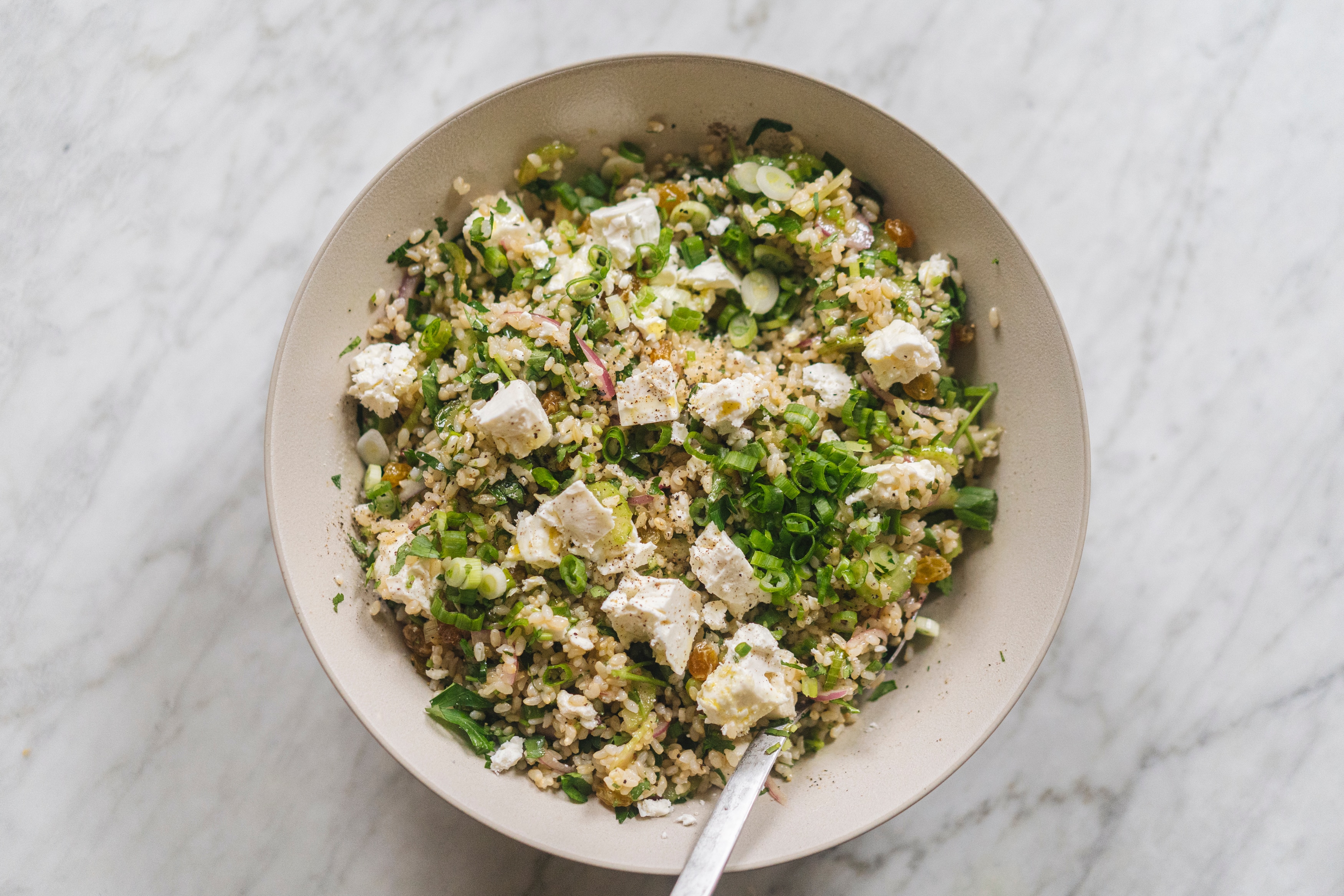 Un tazón blanco lleno de ensalada de arroz integral con queso feta, hierbas picadas y un aderezo rápido de pepinillos.
