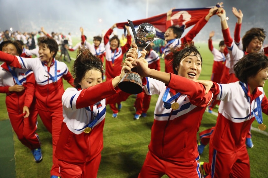 A soccer team wearing red and white hold a trophy and run holding a flag after winning a tournament
