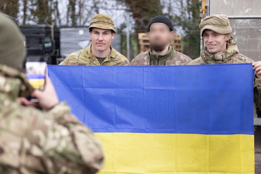 Two Australian soldiers stand either side of a Ukrainian soldier. The three hold a Ukrainian flag