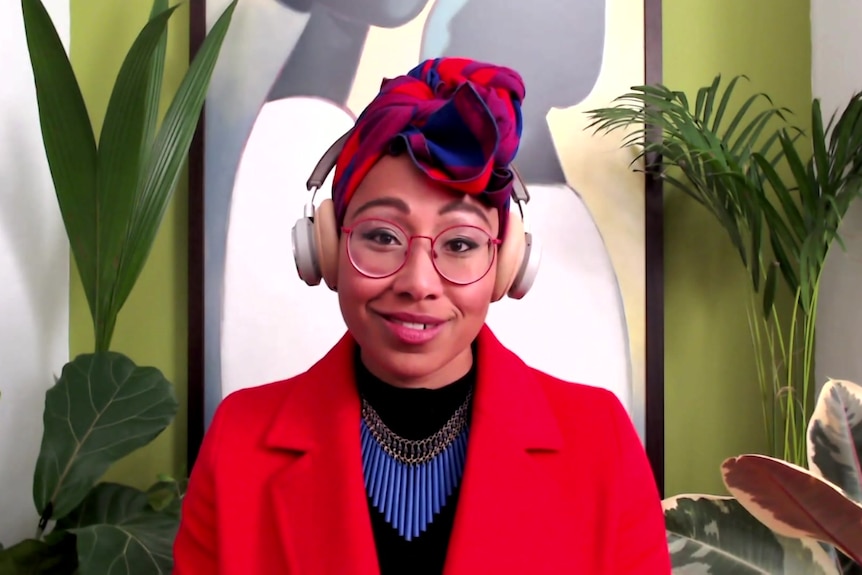 A young Black woman with a red jacket, glasses and head scarf wears headphones and looks directly at the camera
