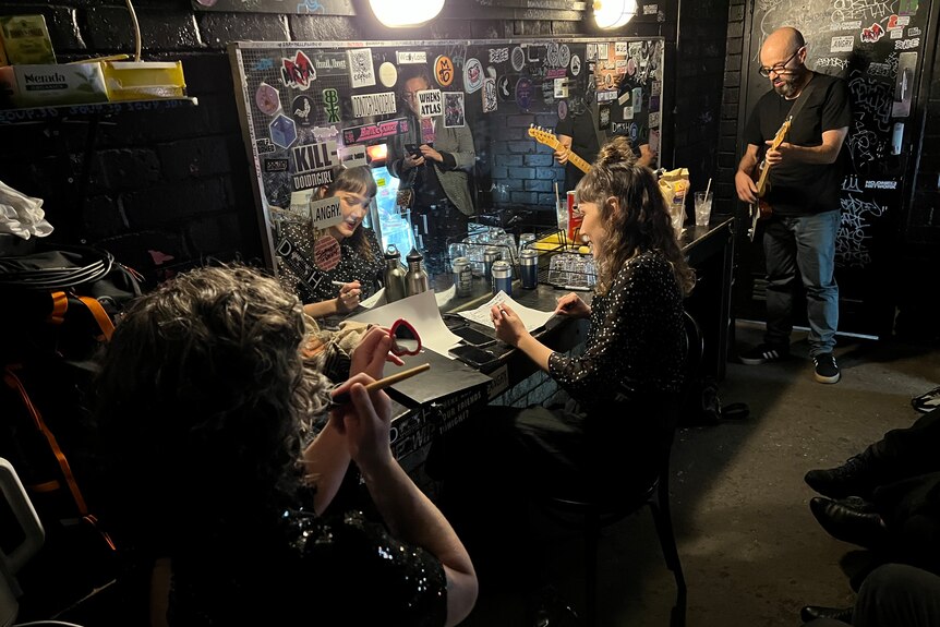 Frente in backstage room: Angie Hart applies make-up, Simon Austin plays guitar, third members writes setlist