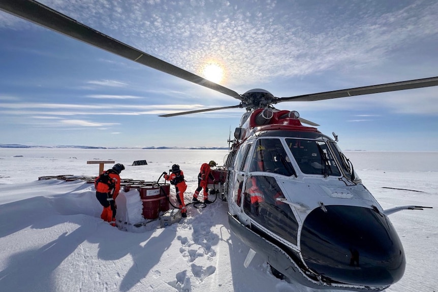 Three people in orange suits stand in the snow beside a rescue helicopter.