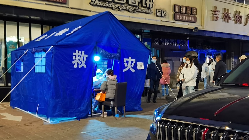 Masked people line up on a street outside a bright blue tent, where COVID tests are being administered
