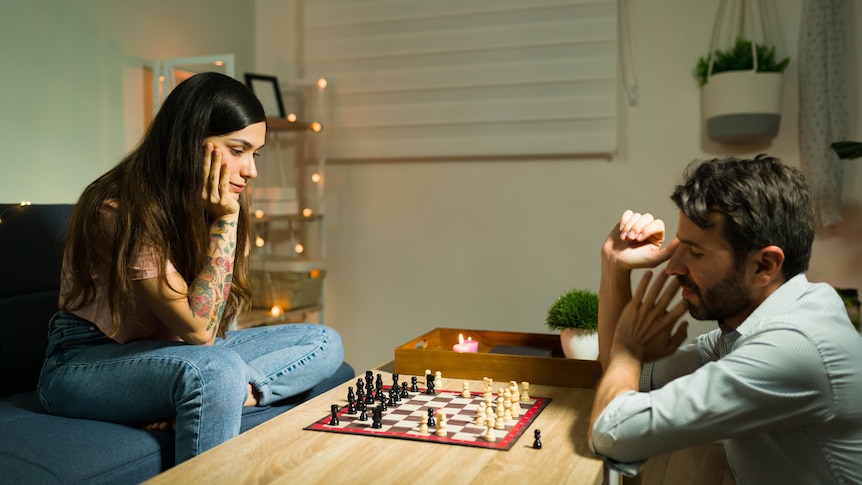 A couple looks serious while playing chess in their living room. The woman sits on the couch while the man sits on the floor.