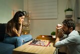 A couple looks serious while playing chess in their living room. The woman sits on the couch while the man sits on the floor.