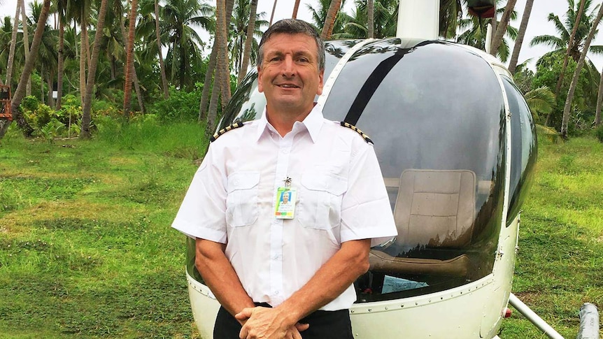 A smiling man stands in front of a helicopter wearing a pilot's uniform. There is palm trees in the far background.