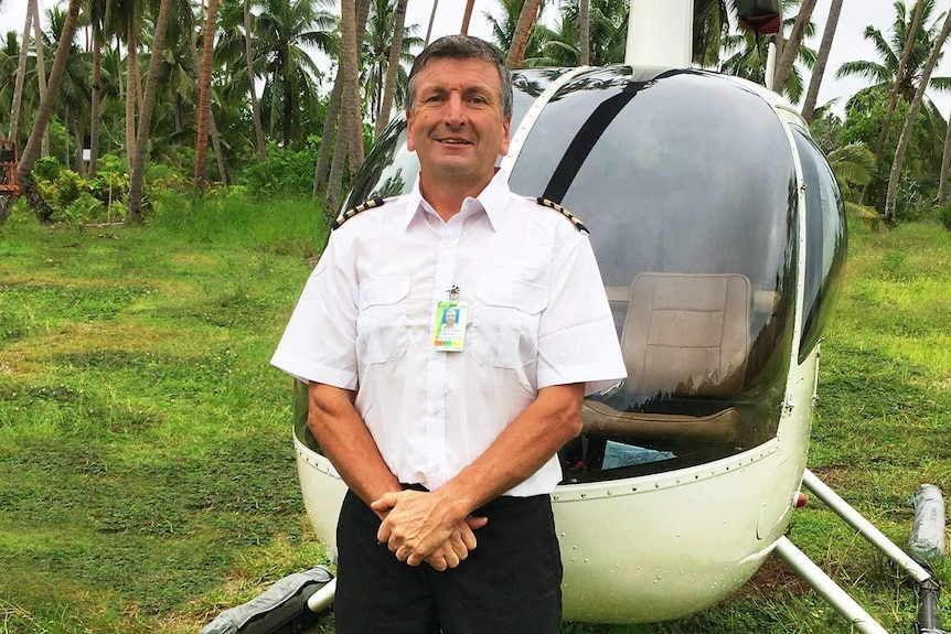 A smiling man stands in front of a helicopter wearing a pilot's uniform. There is palm trees in the far background.