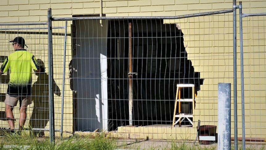 A tradesman stands next to a fenced off smashed wall