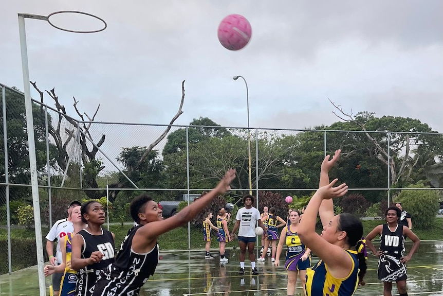 A netball team plays on a wet court, with a player shooting a ball towards a hoop
