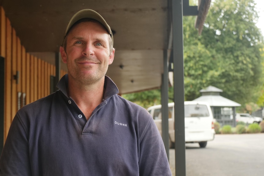 A man wearing a baseball cap smiles at the camera, with caravans behind him.