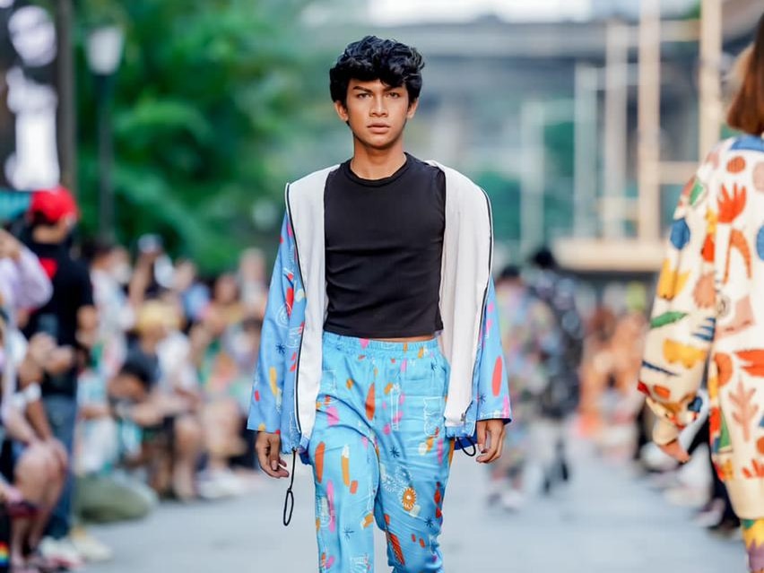 A man walking straight to the camera doing fashion show, wearing black shirt and blue jacket and pants.