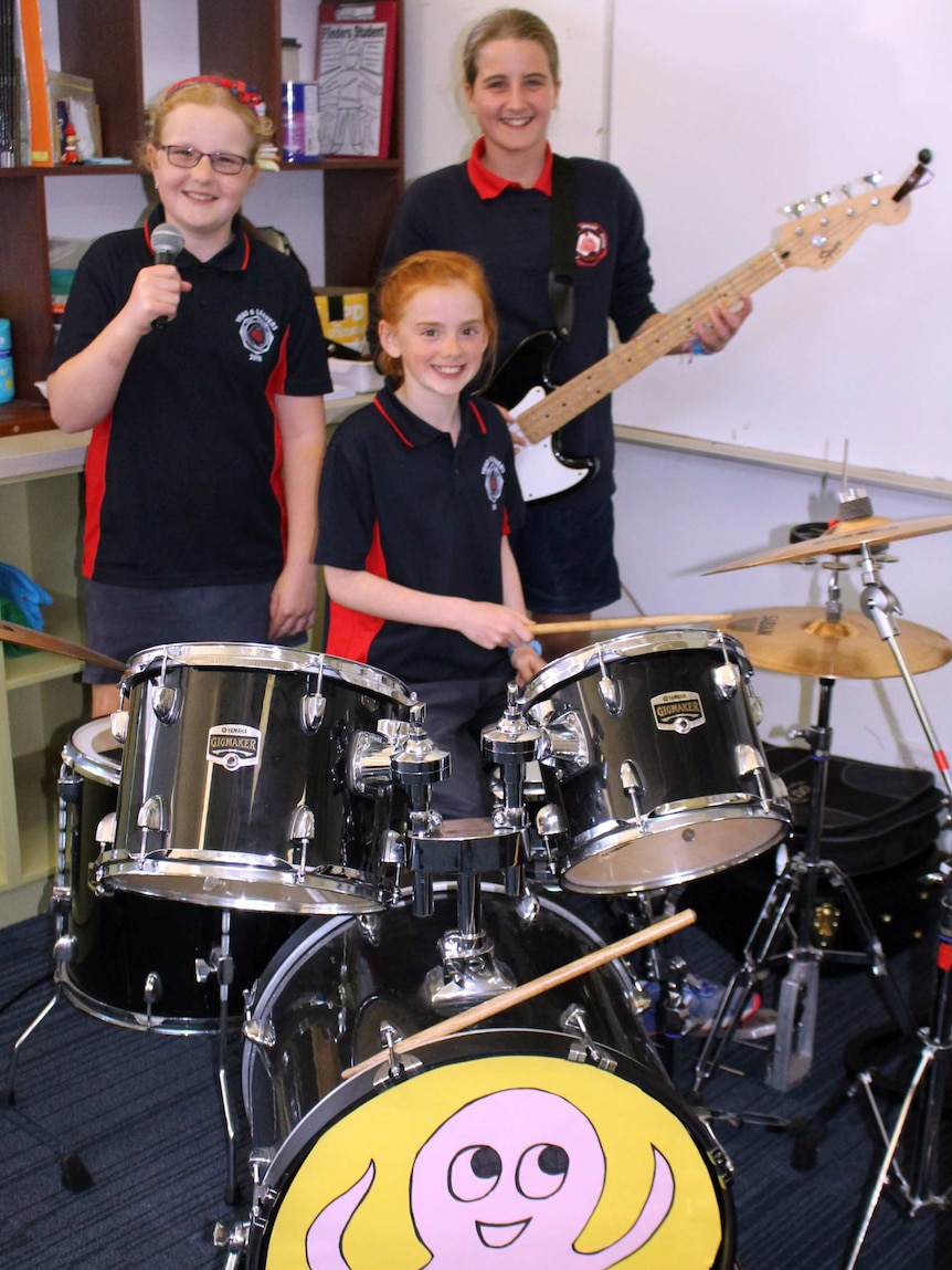Three young girls with a drum kit, bass guitar and microphone in a classroom