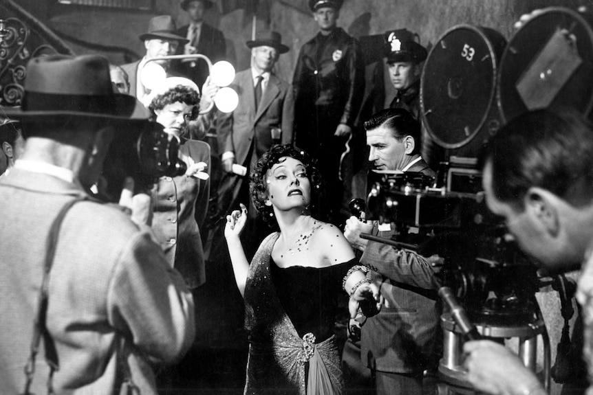 A glamourous woman with a strange look on her face is surrounding by press cameras and police in the 1950s.