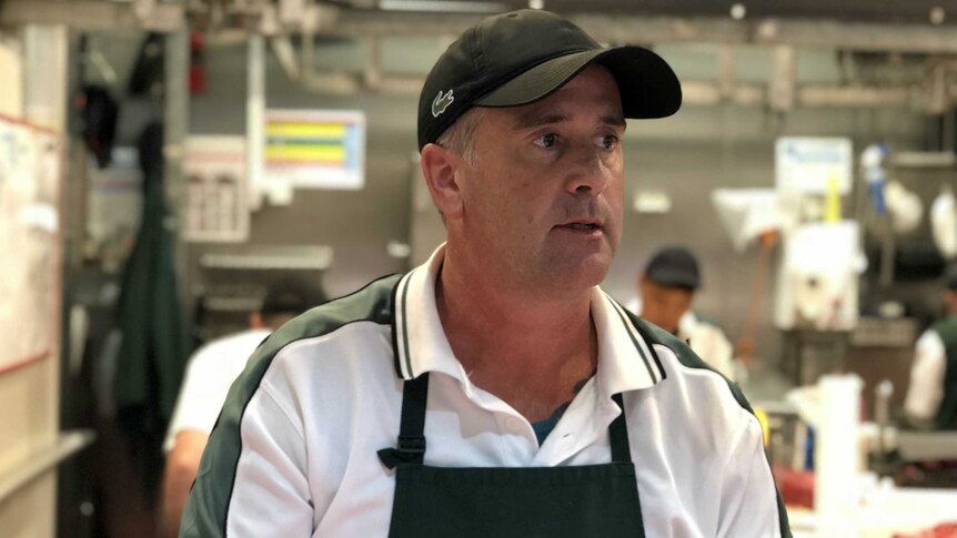 barry stands behind a counter at a butcher shop as workers prepare cuts of meat behind him