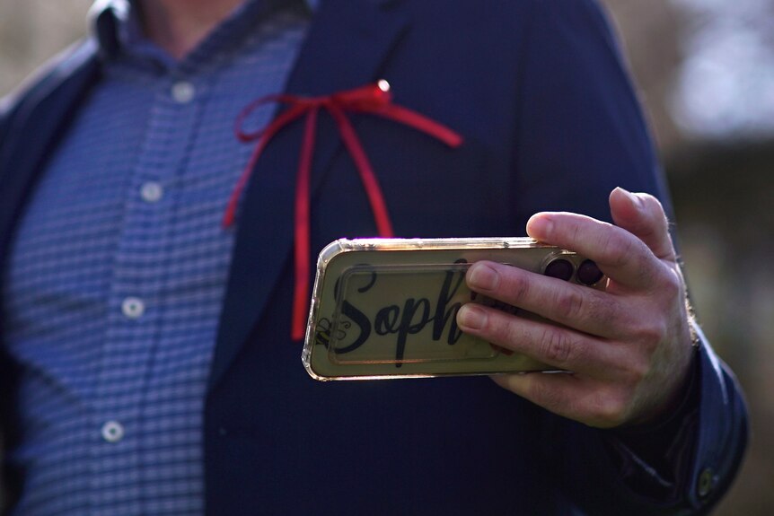 A man's hand holds a phone with a case reading 'Sophia'. He's wearing a suit with a red ribbon