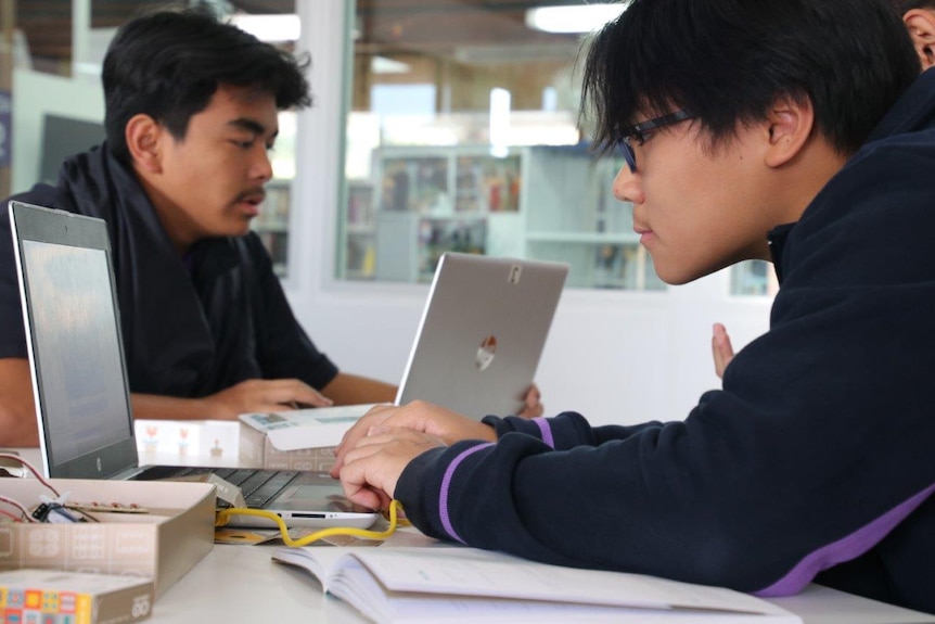 Two boys facing opposite each other working on computers at a school desk.