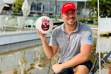 Man with red hat kneeling at boat marina while holding up a volleyball with a hand-print red face.