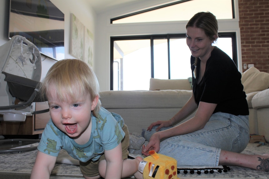 A one-year-old boy crawls towards the camera while his mother smiles on the floor behind him