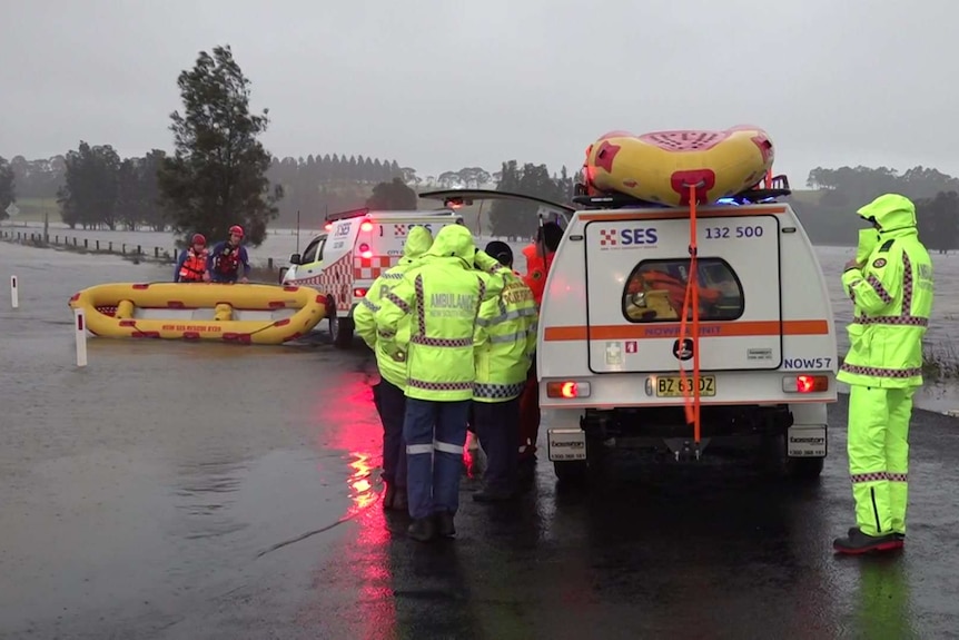 SES vehicles and people wearing high-visibility raincoats stand next to a flooded road.