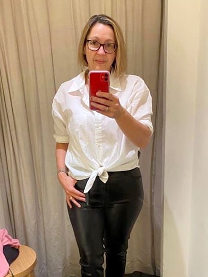 Woman with bobbed hair stands in a dressing room taking a selfie showing white shirt and black leather pants