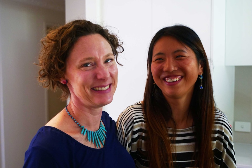 Two women stand in a house, beaming at the camera.