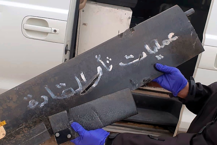 An unseen man with blue gloves holds a black piece of aircraft in his hands with white Arabic writing on it.