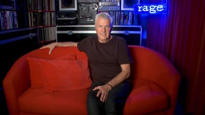 A man in a dark T-shirt sits on a red couch, looking at the camera.