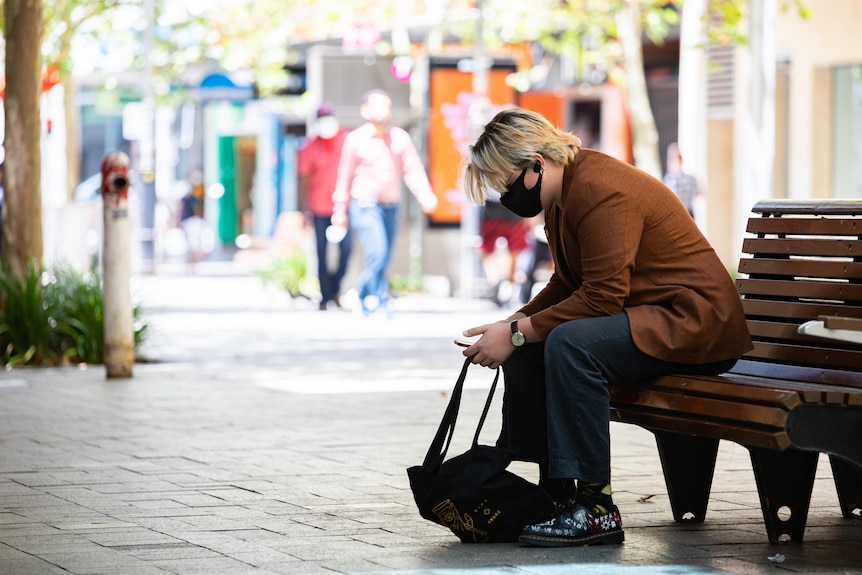 A man sits on a wooden bench, looking at his phone and wearing a mask.