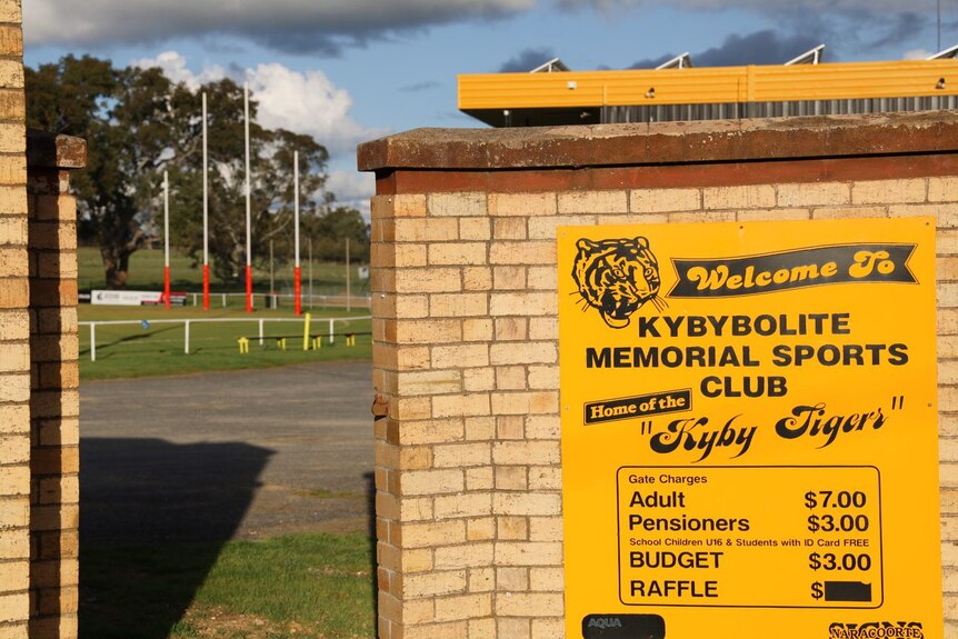 A sign next to a gate opening onto a football oval reads "Welcome to Kybybolite Memorial Sports Club, home of the Kyby Tigers"