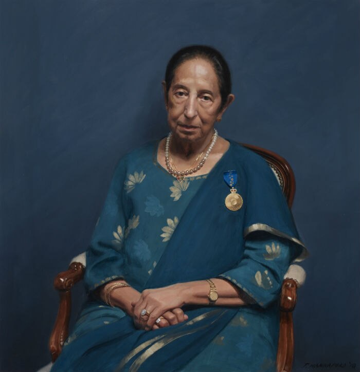 A older woman wearing a blue and aqua sari sits in a wooden arm chair in front of a blue wall.