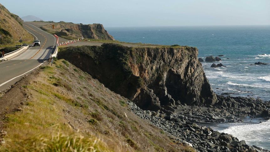 A vehicle drives along a cliffside section of road.