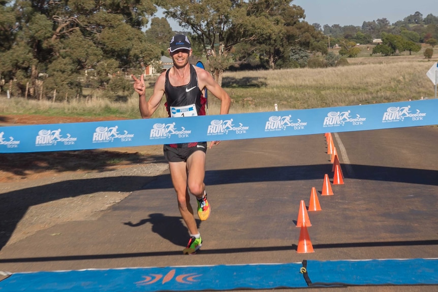 A man with his finger pointed upwards runs through a finish line ribbon on a road in a country area with paddocks behind