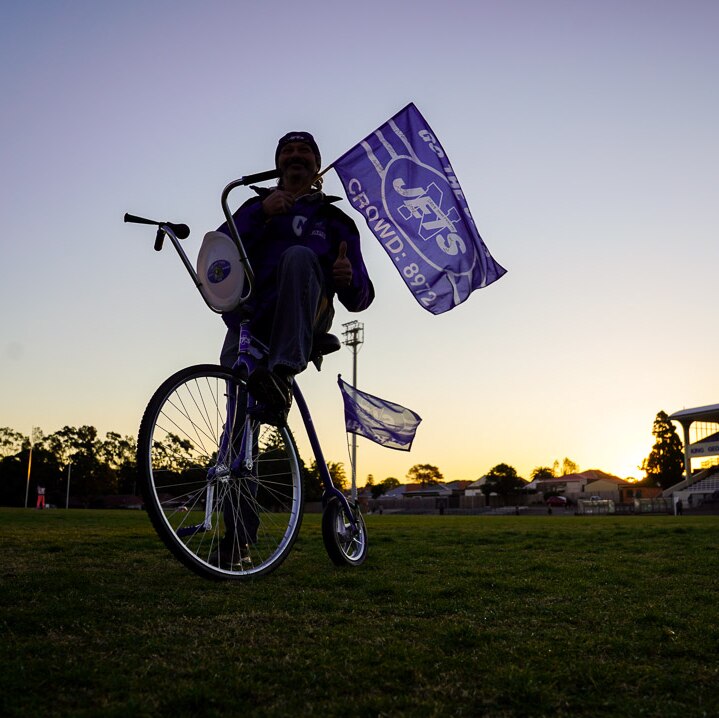 A man stands on Henson Park with a mini-penny-farthing bicycle with the sun and grandstand in the background.
