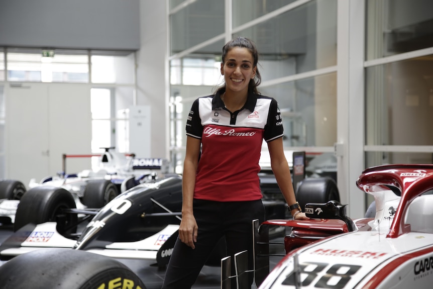 Krystina Emmanouilides stands in front of a formula one car and smiles.