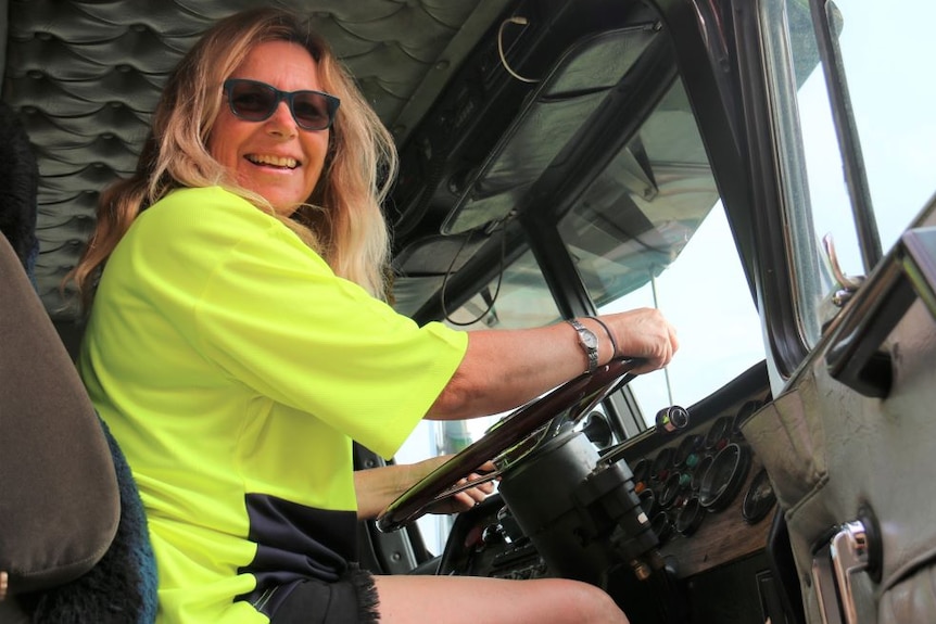 A woman with long blond hair sits in the drivers seat of a truck and smiles