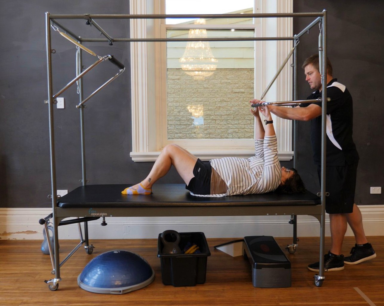 A woman in a black and white stripe top lays on a bed while stretching her arms with a pole with the help of a teacher.