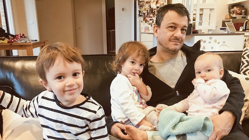 Mark Tamhane sits on a couch holding two young children. A third child, sitting separately on the couch, smiles at the camera.