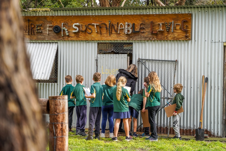 A group of primary kids in green school uniforms stands by a tin shed under a sign reading 'Centre for Sustainable Living'.
