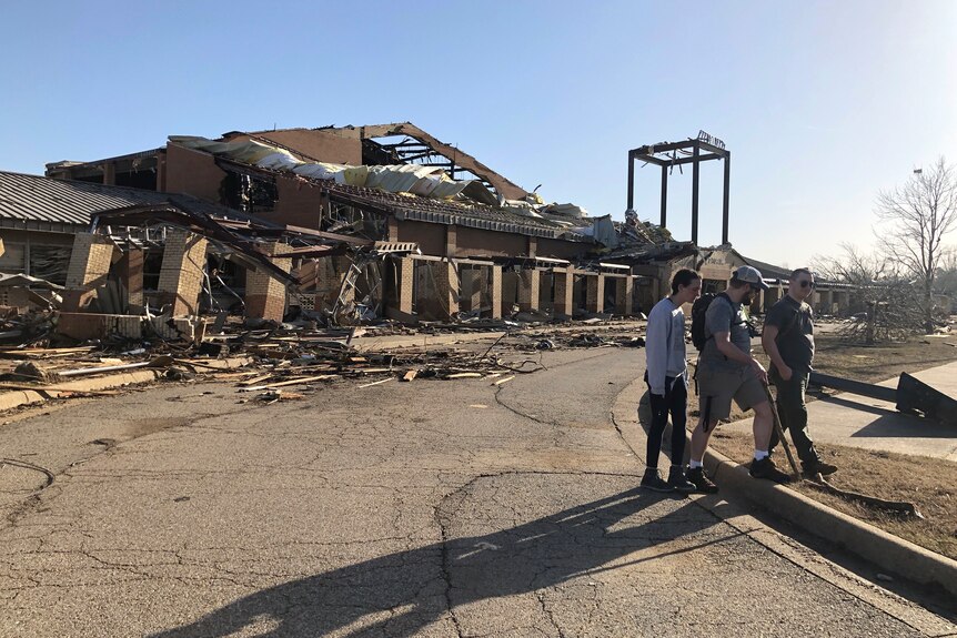 People stand on the road as a building with its roof destroyed can be seen in he background. 
