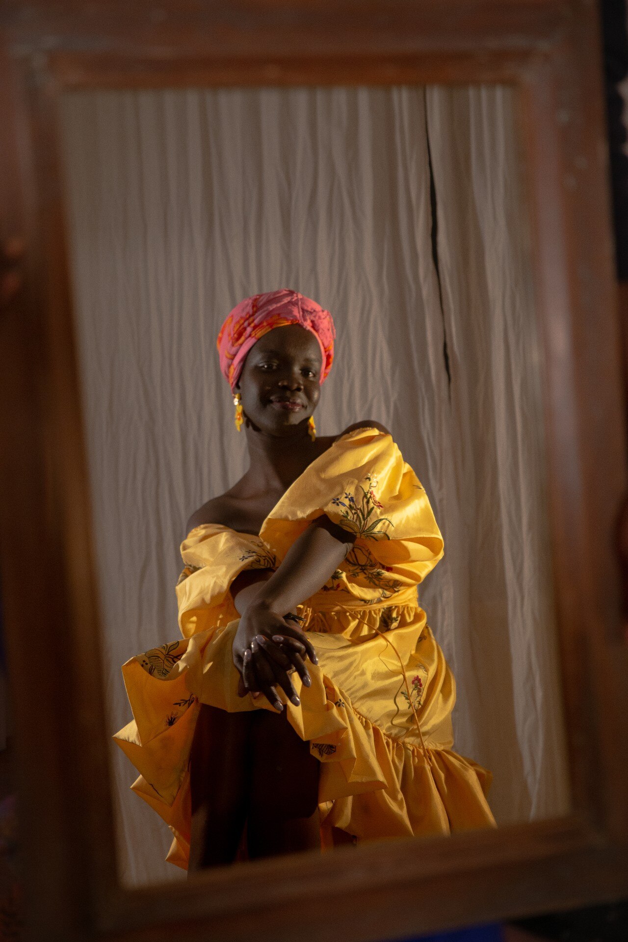 A 20-something African woman poses at the mirror with a smile, wearing a yellow dress and pink turban, her hands on her knees 