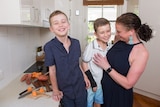 A woman and her two young sons stand in the kitchen together, smiling. 