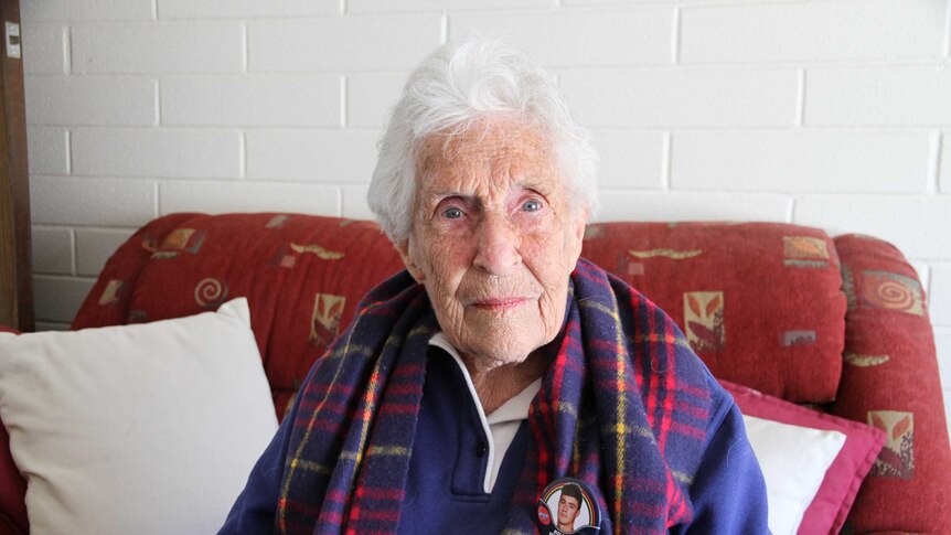 An elderly lady sitting in her armchair wearing and Adelaide Crows scarf and a badge of her grandson Riley Knight