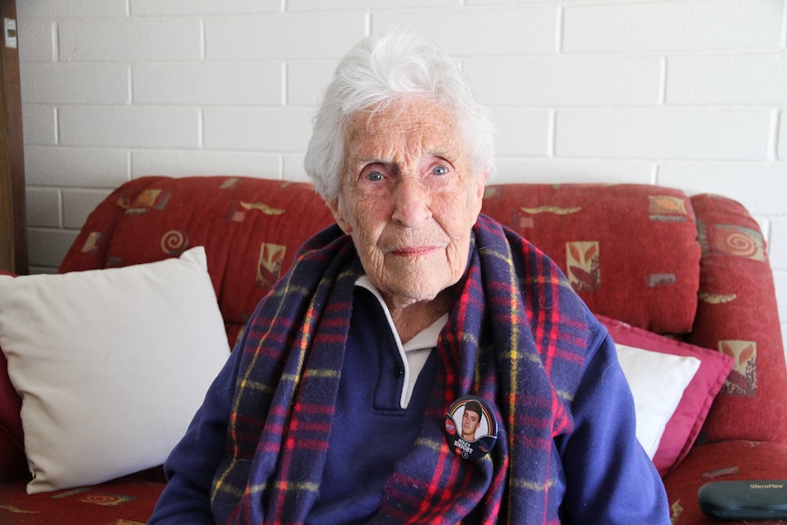 An elderly lady sitting in her armchair wearing and Adelaide Crows scarf and a badge of her grandson Riley Knight