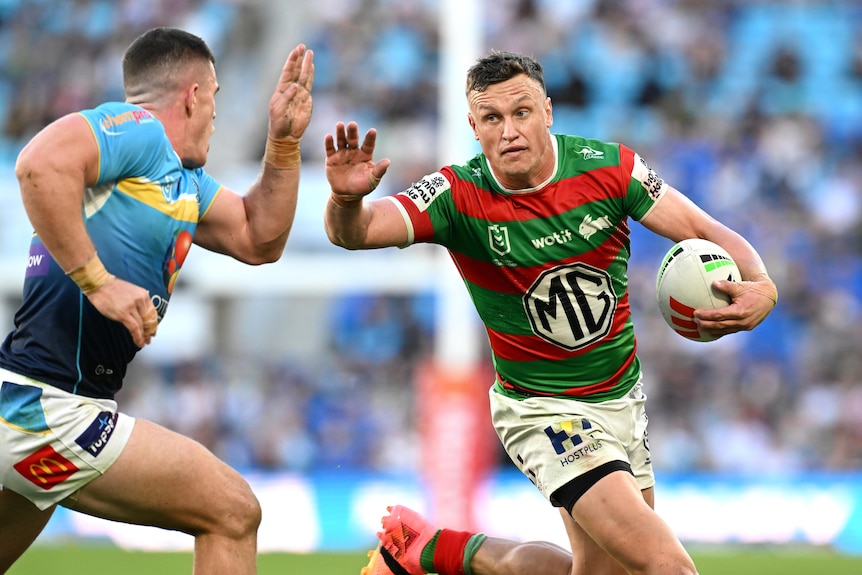 NRL player Jack Wighton of the Rabbitohs fends off a defender while running with the ball in one arm